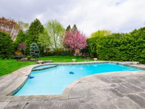 A backyard swimming pool with light blue water, surrounded by grey paving around pool area, lush green lawn, and various trees under a cloudy sky.