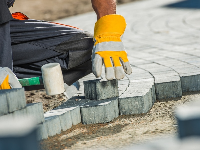 Worker installing natural stone paving blocks with gloves and rubber mallet on outdoor pathway.