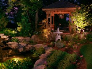 A neat garden at dusk featuring a timber gazebo, soft lighting, stone water feature, and layered plants.