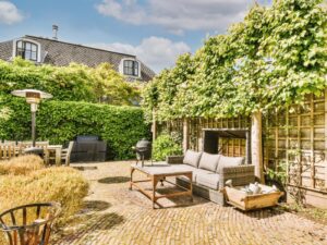 A neat garden courtyard with brick paving, rattan furniture, climbing vines, and trimmed hedges under clear daylight.